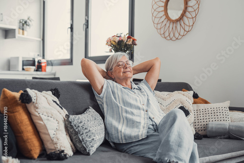 Elderly woman relaxes on a sofa at home, stretching and smiling peacefully. She enjoys a calm and carefree moment, embracing happiness and serenity in her domestic lifestyle.