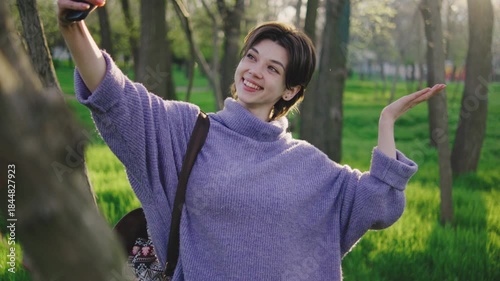Woman takes a selfie in a park during spring with green grass and trees around her under bright sunlight