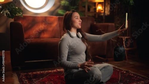 Young woman sitting on the floor at home talks to someone via video call on her smartphone. Cozy interior, casual lifestyle, online communication, remote connection, modern technology