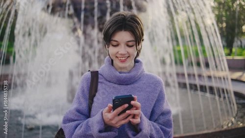 Young woman stands near water fountain in a public park looking at her smartphone while sunlight shines through trees