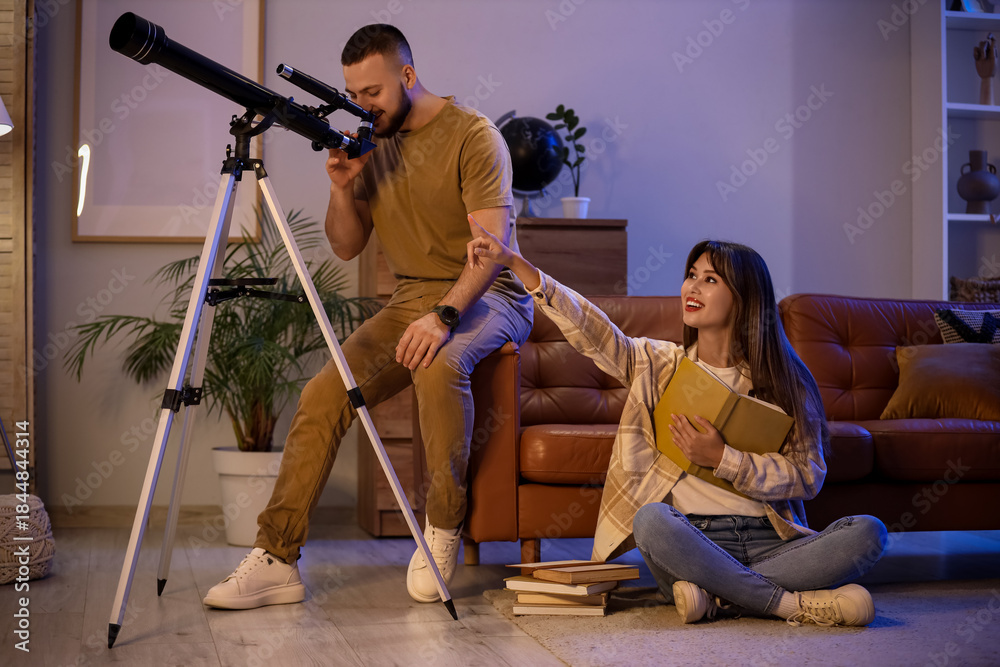 Fototapeta premium Young couple with books looking through telescope in room at night