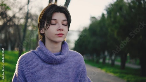 Young woman sits peacefully in a park wearing a purple sweater while meditating on a stone during late afternoon