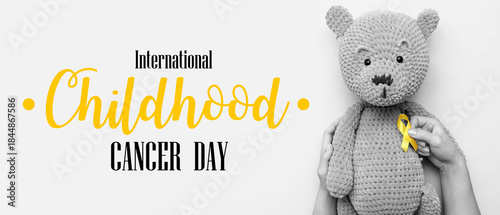 Female hands with knitted toy bear and golden awareness ribbon on grey backgr...