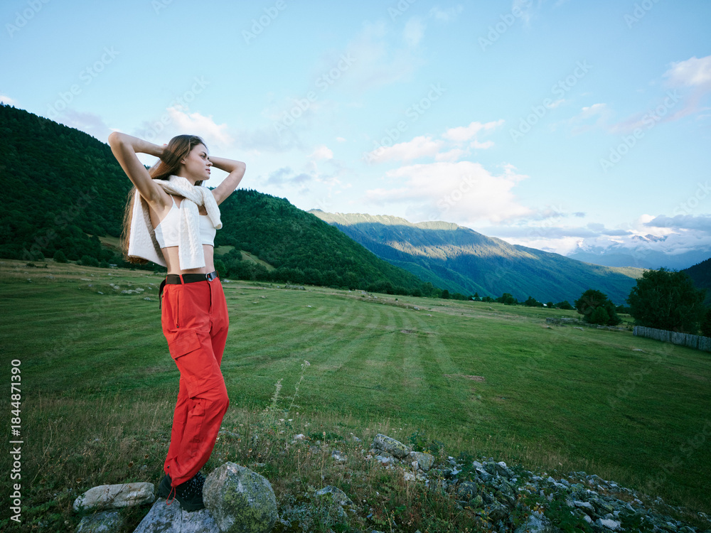 Fototapeta premium Woman in red trousers and scarf stands on rocks in an open field with mountains, capturing a moment of outdoor freedom and scenic landscape