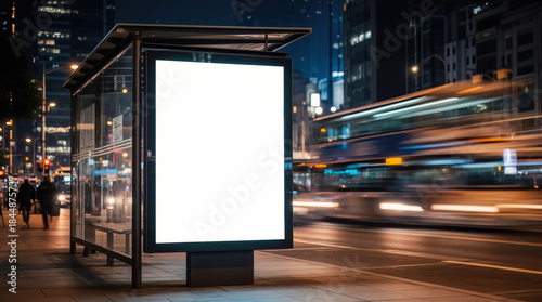 Illuminated blank advertising billboard at a bustling city bus stop at night, capturing the dynamic urban environment with blurred passing buses and vibrant streetlights.