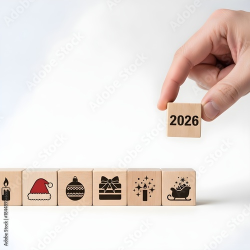 Hand placing wooden block with '2026' on a row of festive Christmas and New Year's icons against a clean white background, symbolizing upcoming celebrations and planning