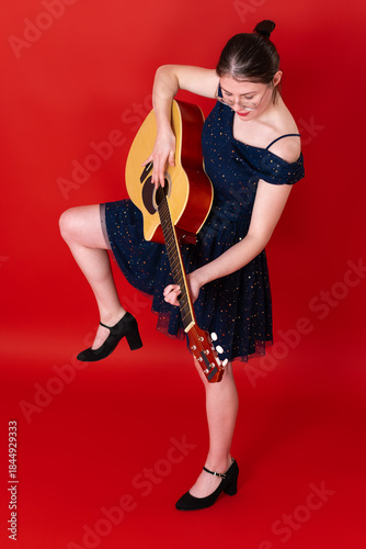 Female guitarist performs with playfulness and technical mastery, standing on one leg, strumming classical guitar with apparent ease, emotional authenticity. Woman wearing glasses, above knee dress