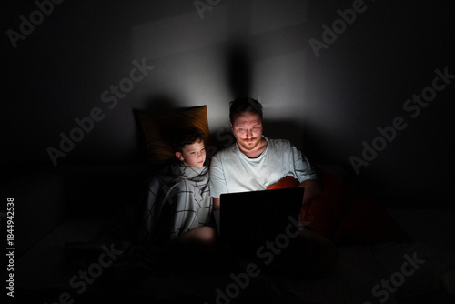 A father and son sit closely on a bed, looking at a laptop screen. The room is dark, with soft light from the laptop illuminating their faces. They are sharing a moment of laughter and joy.