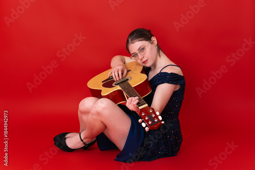 Female musician and guitarist playing guitar while sitting on floor against red background and looking at camera. Young woman singer songwriter wearing eyeglasses, above knee dress, high heeled shoe
