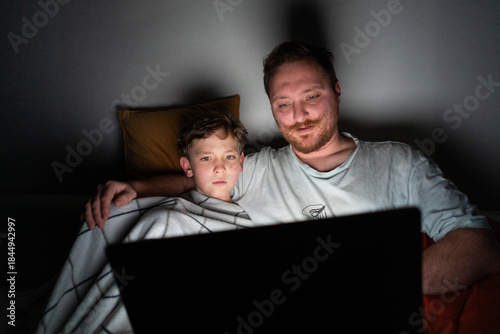 A father and son sit closely on a bed, looking at a laptop screen. The room is dark, with soft light from the laptop illuminating their faces. They are sharing a moment of laughter and joy.