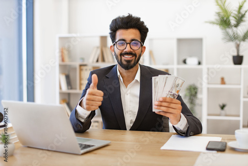 An Indian businessman smiles while holding cash and giving a thumbs up, showing his approval