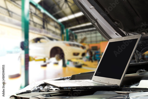 Digital Car Diagnostics, A laptop with blank screen sits on a car engine in a modern auto repair shop, displaying diagnostic data while a vehicle is elevated on a lift for professional service.