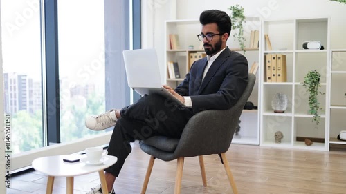 A handsome Arab businessman sits in a modern office chair, working on his laptop, looking thoughtful