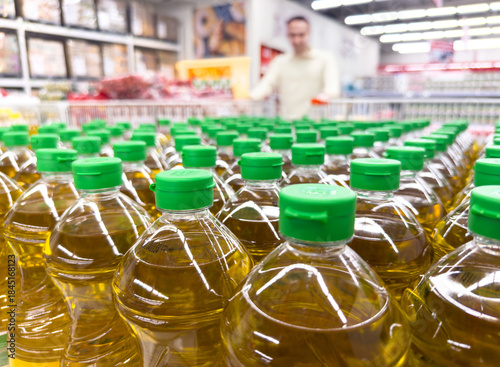 low-angle shot of many clear plastic bottles of golden cooking oil with green caps lined up on a grocery store shelf, with a male customer blurred in the background, symbolizing healthy eating 