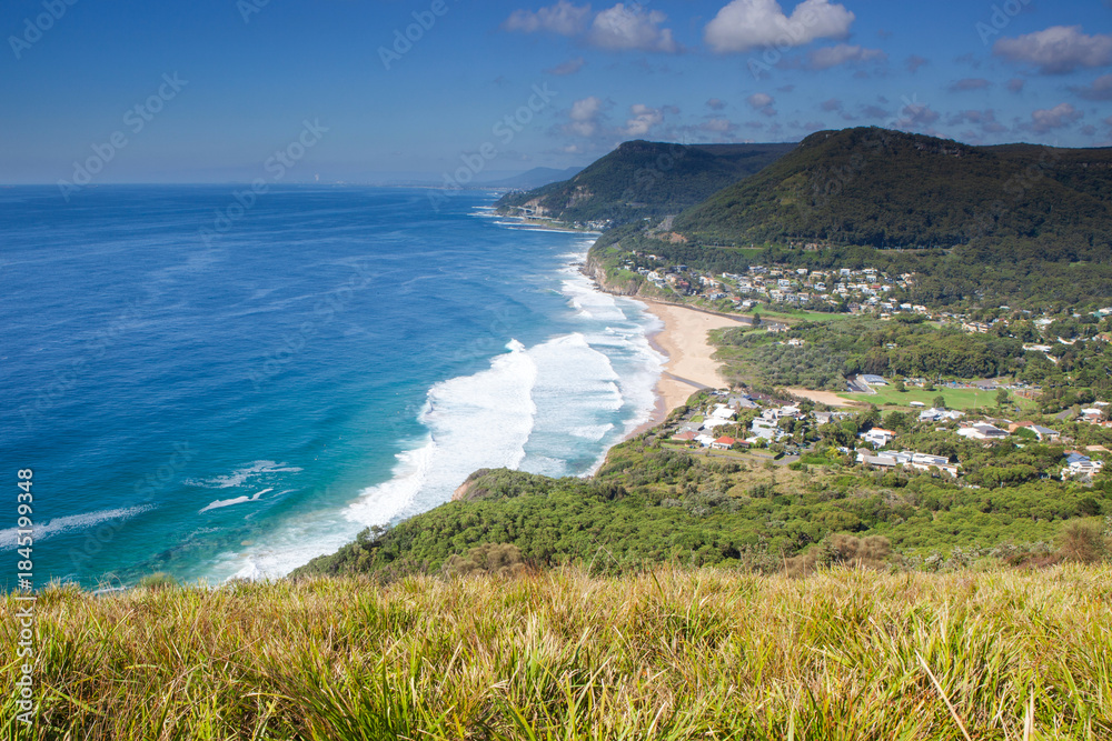 Naklejka premium The iconic view looking south to Stanwell Park beach and the famous Sea Cliff Bridge from the Illawarra's Bald Hill Lookout. Captured on a blue sky sunny day from the grassy knoll.
