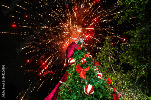 Festive Christmas tree decorated with red bows and ornaments under night sky with bright fireworks explosion above, holiday celebration outdoors, New Year atmosphere, winter festival mood