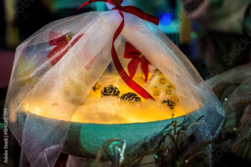 Glowing decorative light arrangement wrapped in sheer fabric with red ribbon, pine cones, cozy festive atmosphere, magical illumination, holiday decor concept, warm mood, creative still life