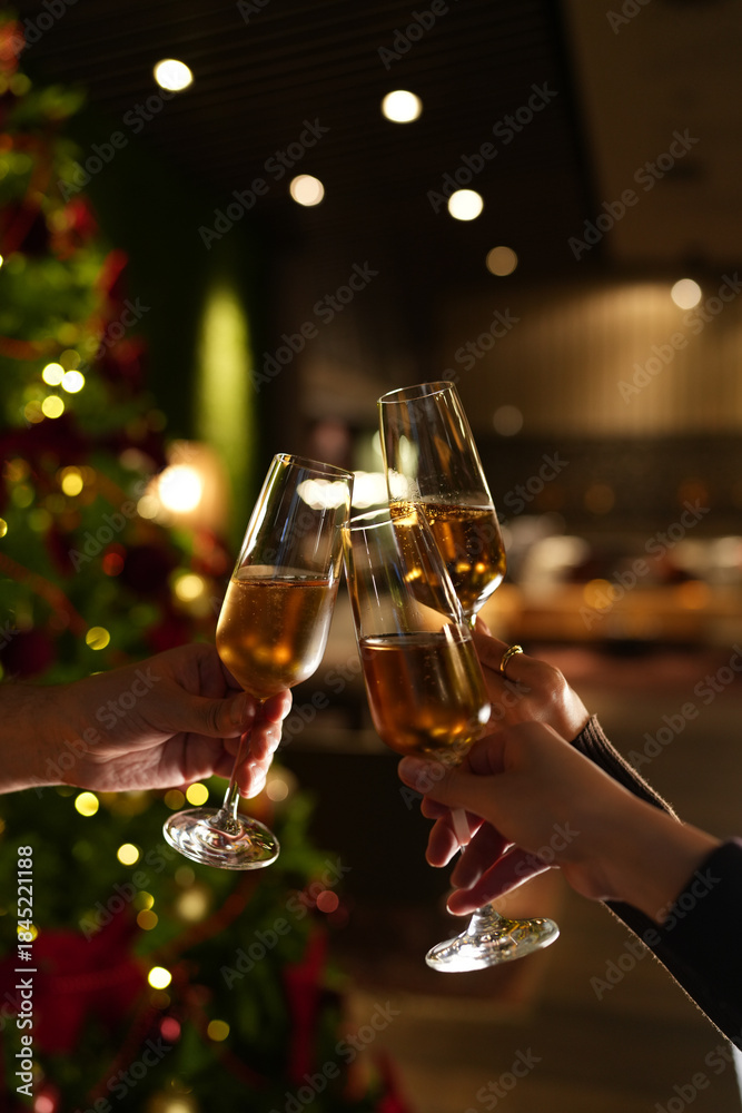 Obraz premium Hands raising glasses of champagne for a festive toast in front of a decorated Christmas tree. Warm holiday atmosphere, bokeh lights, celebration mood and New Year party vibes.
