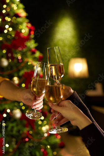 Hands raising glasses of champagne for a festive toast in front of a decorated Christmas tree. Warm holiday atmosphere, bokeh lights, celebration mood and New Year party vibes.