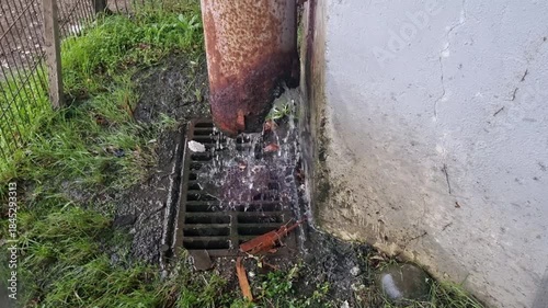 Closeup of a rusty metal downspout channeling rainwater into a grimy urban storm drain. Water flowing down the weathered pipe