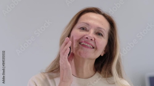 A woman is smiling and applying makeup. She is wearing a white shirt. She is using a brush to apply makeup