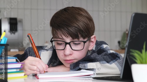 A boy is writing in a notebook. He is wearing glasses. The notebook is on a table