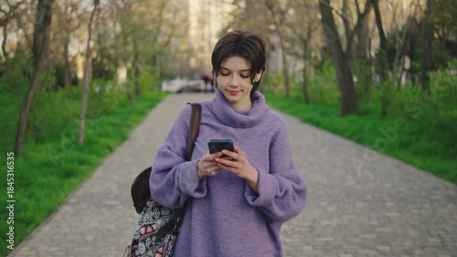 Young woman walks on a paved path in a park while using a smartphone during afternoon light