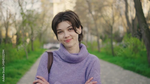 Young woman stands on path in green park setting during daytime with city buildings visible in background, smiling softly and crossing arms