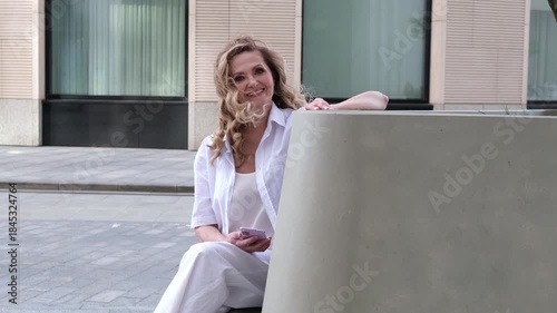 A woman sitting on a bench with her hand on her hip. She is wearing a white shirt and white pants