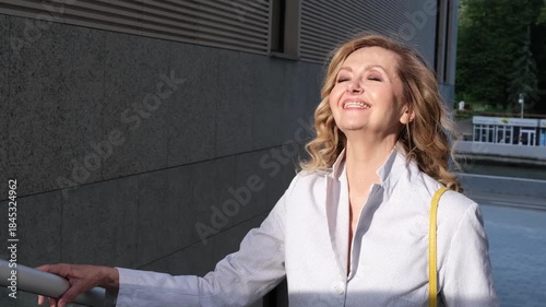 A woman wearing a white shirt and yellow purse is standing on a ledge. She is smiling and looking at the camera