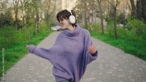 Young woman dances joyfully on a path in a park while wearing headphones during daytime surrounded by trees and greenery