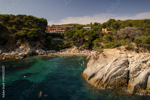Aerial view of azure waters meeting golden sands beneath verdant cliffs, where swimmers and sunbathers revel in the summer glow, Platja d'Aro, Catalonia, Spain.