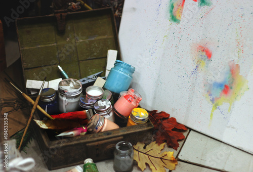Vintage art supplies in an old wooden box: colorful jars of paint, brushes, and a dry leaf next to a messy canvas, symbolizing creative process and artistic inspiration