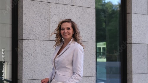 A woman with long hair is standing in front of a building. She is wearing a white jacket and white pants
