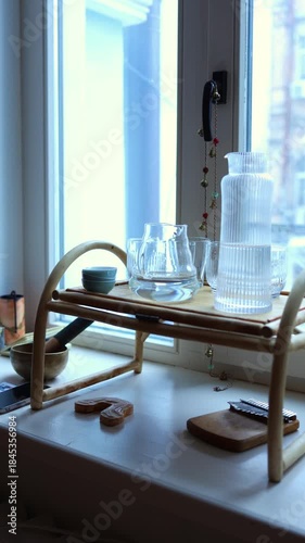 Still life featuring minimalist tea ceremony accessories, including a wooden stand, clear glass teapot cups, small bowls, a singing bowl, and meditation chimes/beads, placed on a sunlit windowsill.