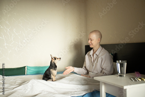 Adult female cancer patient in pajamas resting in bed with tiny dog, glass of water and pills on bedside table, home treatment