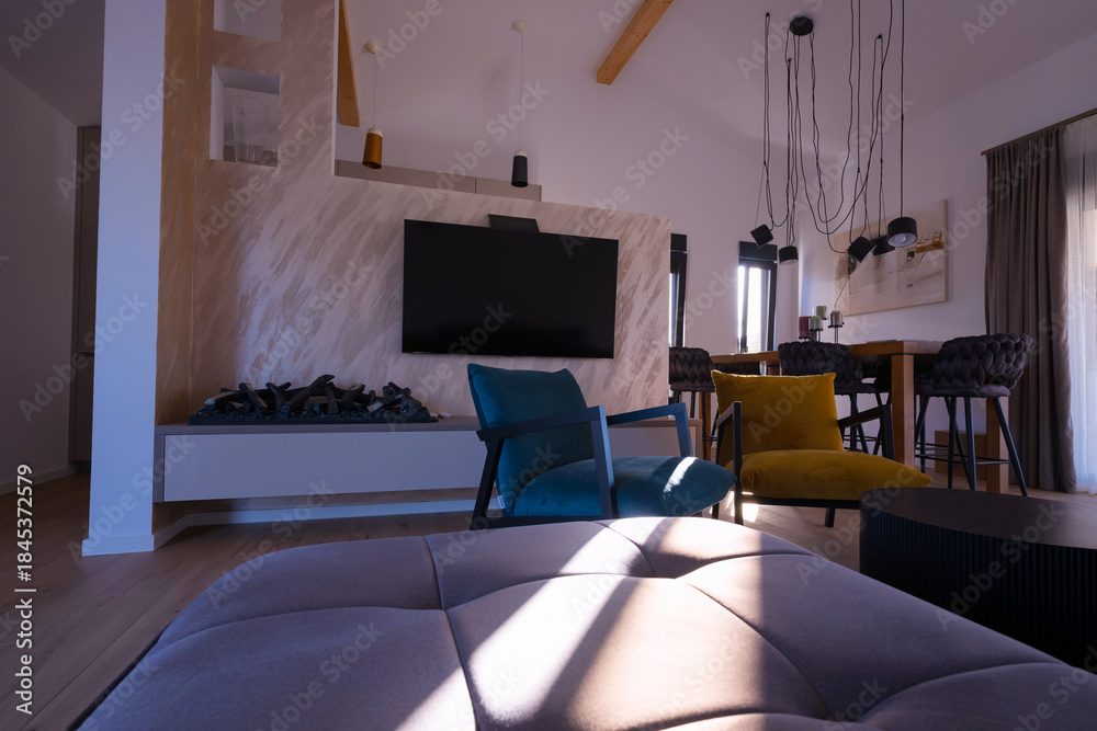 Obraz premium Modern open-plan living and dining area with an integrated kitchen, featuring a comfortable sofa, stylish armchairs, and a bar-height dining table. Wooden floors and ceiling beams enhance this