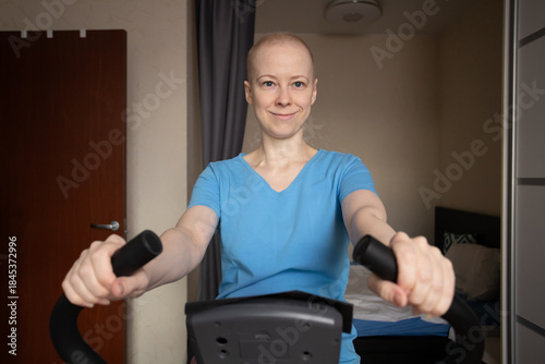 Cancer patient works out at home on an exercise bike, woman after chemotherapy training in bedroom, indoor cardio workout for oncology rehabilitation, home fitness and recovery