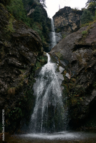 Water cascades down steep rocks surrounded by dense greenery in a forest. The scene takes place on a cloudy day, adding a moody atmosphere to the setting