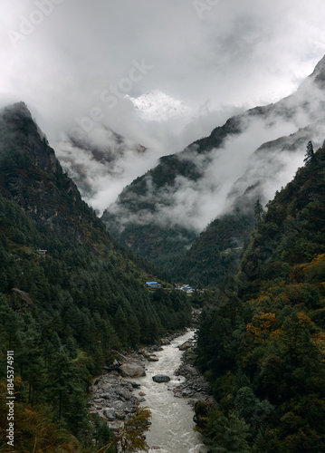 Clouds cover the mountain peaks while a river flows through a green valley. Trees line the banks and fog rolls over the hills. The scene shows nature's beauty in a secluded area