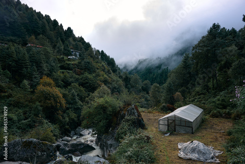 A camp is set up in a valley surrounded by trees and hills. A stream flows nearby, and clouds linger above the mountain tops, creating a mix of nature and shelter