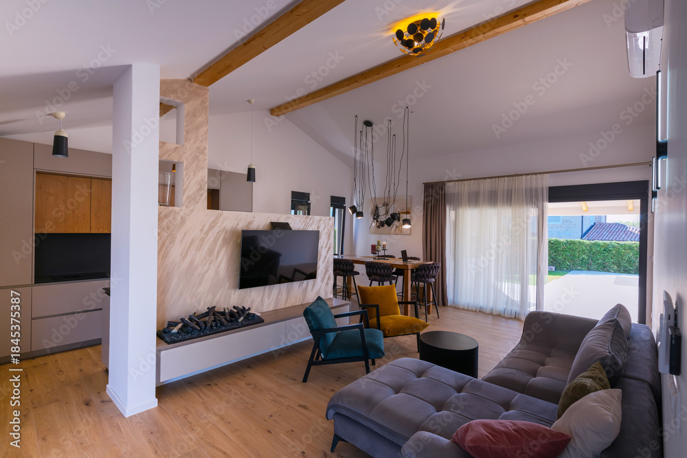 Obraz premium Modern open-plan living and dining area with an integrated kitchen, featuring a comfortable sofa, stylish armchairs, and a bar-height dining table. Wooden floors and ceiling beams enhance this