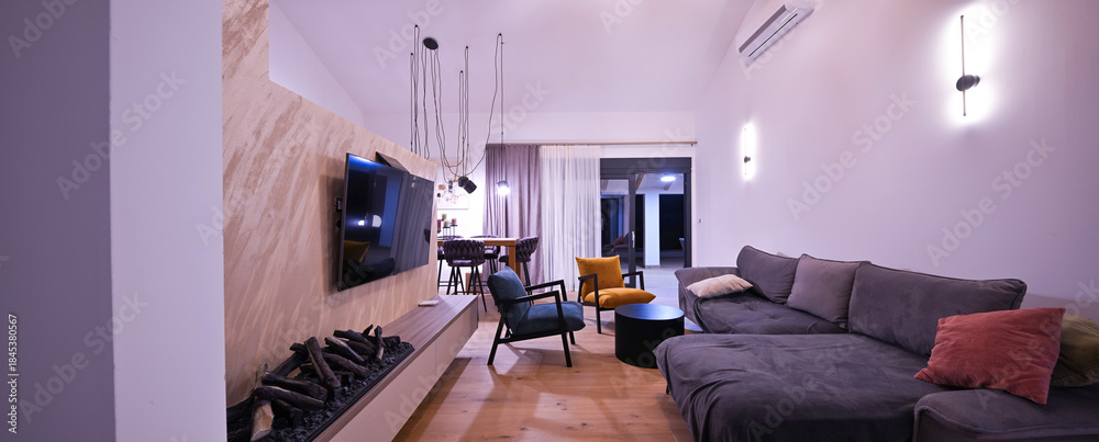 Obraz premium Modern open-plan living and dining area with an integrated kitchen, featuring a comfortable sofa, stylish armchairs, and a bar-height dining table. Wooden floors and ceiling beams enhance this