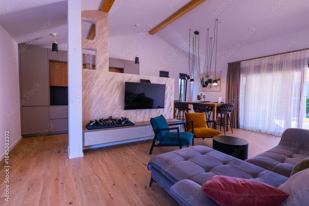 Obraz premium Modern open-plan living and dining area with an integrated kitchen, featuring a comfortable sofa, stylish armchairs, and a bar-height dining table. Wooden floors and ceiling beams enhance this