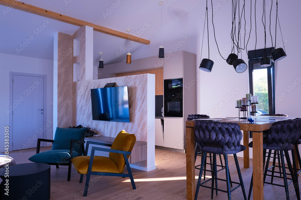 Fototapeta premium Modern open-plan living and dining area with an integrated kitchen, featuring a comfortable sofa, stylish armchairs, and a bar-height dining table. Wooden floors and ceiling beams enhance this