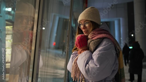 Lovely young lady dressed in cozy winter attire, gazing at a bright store display on the snow-covered city streets at night, basking in the joyful ambiance of the holiday season during christmas