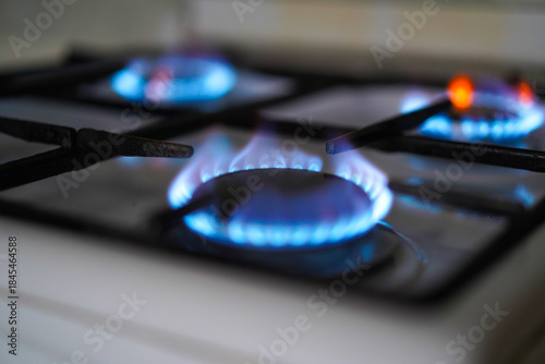 Close-up of four gas stove burners lit, showing hot blue and orange flames on a dark, reflective surface.
