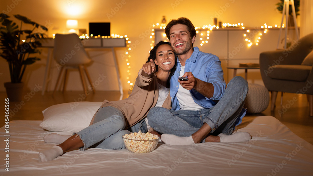 Obraz premium Millennial couple is having fun watching a movie in their bedroom. They are relaxed and pointing at the TV screen while sitting on a comfortable bed with a bowl of popcorn.