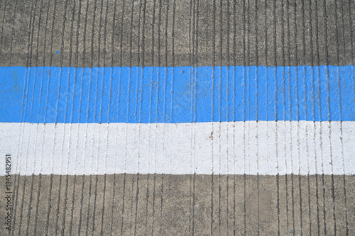 A close-up photograph of a concrete road surface featuring a vertical ridged texture.Blue and white strip are painted on the concrete road.