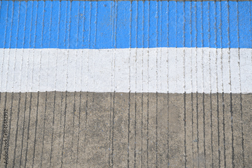 A close-up photograph of a concrete road surface featuring a vertical ridged texture.Blue and white strip are painted on the concrete road.